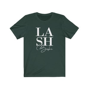 Pu&ograve; includere: T-shirt verde scuro con la scritta bianca "LASH Babe". La parola "LASH" &egrave; sovrapposta a "Babe", con "SH" in mezzo. La parola "Babe" &egrave; in corsivo.