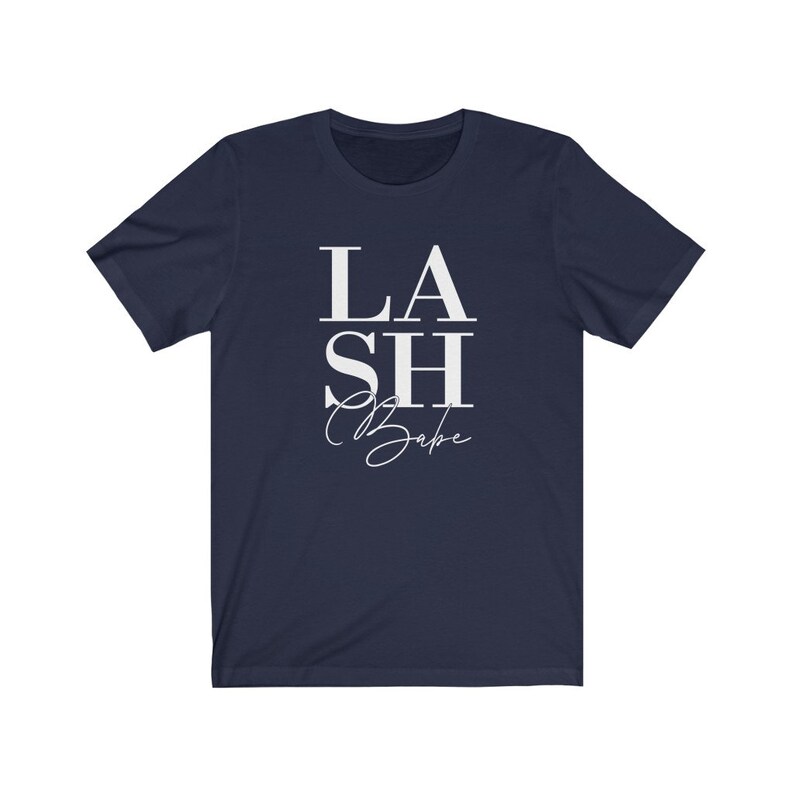 Pu&ograve; includere: T-shirt blu navy con la scritta bianca "LASH Babe". La parola "LASH" &egrave; sovrapposta a "SH", con "Babe" in un carattere corsivo sottostante. La maglietta &egrave; a girocollo e maniche corte.