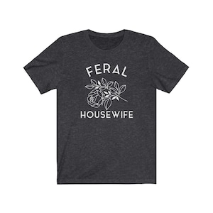 May include: A dark heather grey t-shirt with the text "FERAL HOUSEWIFE" in white lettering. A stylized white rose with leaves is above the text.