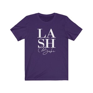 Pu&ograve; includere: Una maglietta viola con la scritta bianca "LASH Babe". La parola "LASH" &egrave; in grassetto, con un carattere sans-serif, mentre "Babe" &egrave; in corsivo. La maglietta ha maniche corte e girocollo.