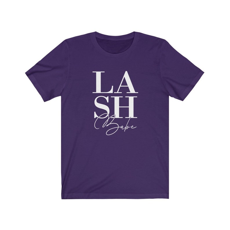 Pu&ograve; includere: Una maglietta viola con la scritta bianca "LASH Babe". La parola "LASH" &egrave; in grassetto, con un carattere sans-serif, mentre "Babe" &egrave; in corsivo. La maglietta ha maniche corte e girocollo.