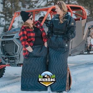 May include: Two people wearing black quilted skirts with a chevron pattern stand in front of a red and black off-road vehicle. The text "KickSac" is visible on the bottom of the image.