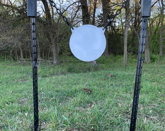 May include: A white round shooting target hanging from a black metal frame with two black metal posts. The target is hanging from a chain and is in a grassy field.