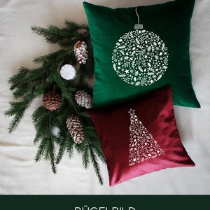 Può includere: Due cuscini di velluto, uno verde smeraldo e uno bordeaux, con disegni bianchi per le vacanze. Il cuscino verde presenta un design di ornamento dettagliato e il cuscino bordeaux ha un design ad albero di Natale. Un rametto di pino e pigne sono sullo sfondo.