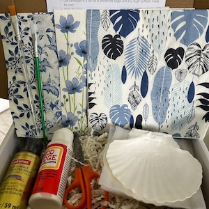 Peut inclure: Un kit de bricolage avec des instructions de découpage comprend de la peinture, du Mod Podge, des ciseaux, des pinceaux et du papier décoratif avec des motifs floraux et de feuilles bleues. Un coquillage blanc est également inclus.