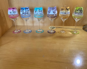 Puede incluir: Seis copas de vino con diseños pintados y refranes humorísticos. Los refranes incluyen "Menos pensar, más vino", "Seguridad primero, imprima con una enfermera", "¡Brindo por eso!", "Hazlo con vino, demasiado", "Bebe feliz, está bien", y "Bebo 1/2 botella periódicamente".