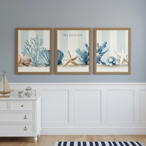 May include: Three framed coastal art prints featuring seashells, coral, and starfish on a sandy beach with blue and white striped backgrounds. The middle print has the name "MEREDITH" in blue text. The frames are a light brown color.
