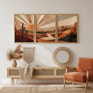 Könnte beinhalten: Drei gerahmte Kunstdrucke, die eine Wüstenlandschaft mit Sonnenuntergang und Kakteen zeigen. Das Kunstwerk verwendet warme Brauntöne, Orange und Beige. Die Drucke werden über einem Holzschrank mit gewebter Front, einem Makramee-Wandbehang und einem runden Spiegel ausgestellt.