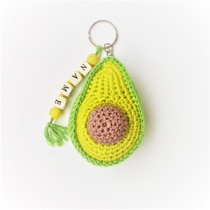 Puede incluir: Un llavero de aguacate tejido a crochet con un diseño de aguacate verde y amarillo y un hueso marrón. El llavero tiene una etiqueta de nombre con cuentas que dice "NAME" en letras blancas.