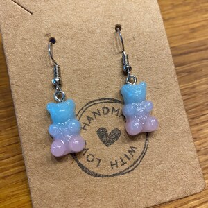 Könnte beinhalten: Ein Paar silberne Ohrhänger mit rosa und blauen Gummibärchen-Anhängern. Die Anhänger sind durchscheinend und haben eine glitzernde Oberfläche. Die Ohrringe sind handgefertigt mit Liebe.
