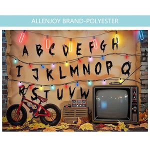 May include: A backdrop featuring a wall with a string of colorful lights and the alphabet in black letters. A red bicycle, a wooden crate with a radio, and a vintage television set are positioned in front of the wall on a floor with autumn leaves.