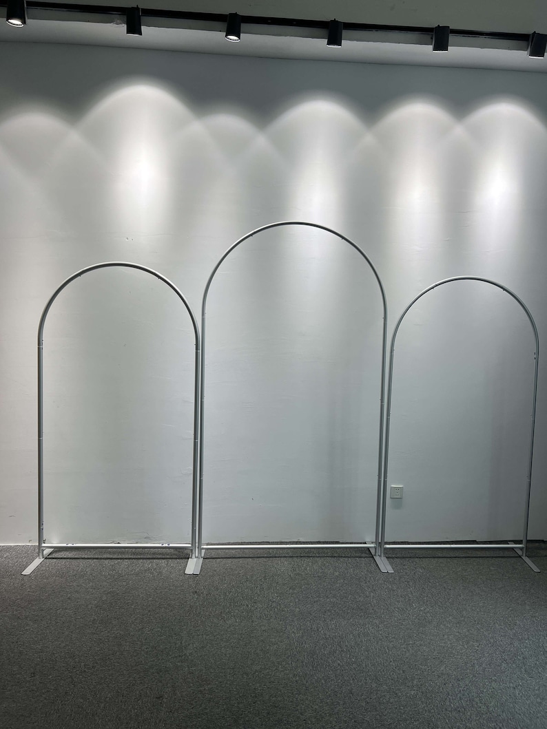 May include: Three silver metal arch backdrops for photography or event decor. The arches are freestanding and have a simple, modern design.