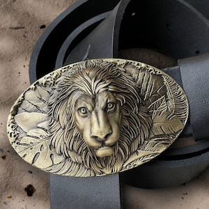 Può includere: Cintura in pelle nera con fibbia ovale in ottone con un design dettagliato a testa di leone. La fibbia è circondata da foglie, creando un contrasto sorprendente. La cintura è larga circa 3,8 cm.