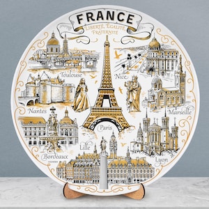 Op de afbeelding: Een decoratieve witte plaat met illustraties van Franse bezienswaardigheden, waaronder de Eiffeltoren, en stadsnamen als Parijs, Nice en Lyon. De plaat heeft een gouden rand en het woord "FRANCE" bovenaan.
