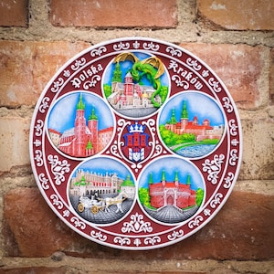 May include: A round, decorative plate with a red, white and blue design. The plate features images of buildings and a dragon, with the words "Polska" and "Krakow" in the top corners.