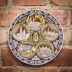 May include: A round, decorative plate with a gold and brown design. The plate features images of buildings and a dragon, with the words "Polska" and "Krakow" in the top corners.