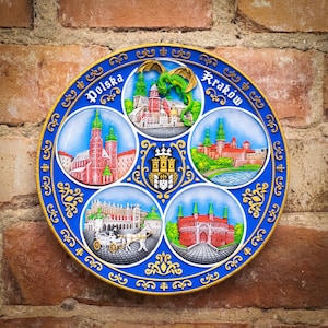 May include: A blue and gold round ceramic plate featuring five images of Polish landmarks. The plate displays the words "Polska" and "Krakow" in gold lettering. The images include a dragon, a castle, a church, and a city square.