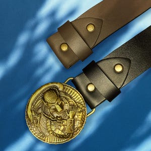 Close-up of brass buckle with Predator warrior engraving on leather belt.