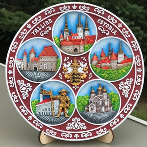 May include: A round, red and white ceramic plate with a decorative border. The plate features five images of buildings in Tallinn, Estonia, including the Alexander Nevsky Cathedral, the Tallinn Town Hall, and the St. Nicholas Church. The plate also includes the text "Tallinn" and "Estonia".