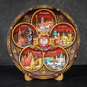 May include: A decorative brown plate with embossed images of Polish cities: Warsaw, Gdańsk, Kraków, Wrocław, and Poznań. The plate has a gold border and a central emblem with the word "POLSKA". It is displayed on a wooden stand.