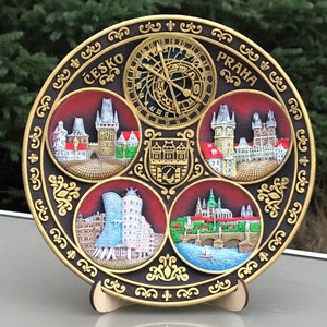 May include: A round, black and gold decorative plate featuring a clock face and four scenes of Prague, Czech Republic. The plate reads "Česko" and "Praha" in gold lettering.