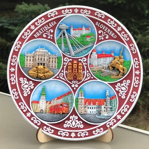 May include: A red and white ceramic plate with a decorative border and five circular images of landmarks in Bratislava, Slovakia. The plate features the text "Bratislava" and "Slovensko".