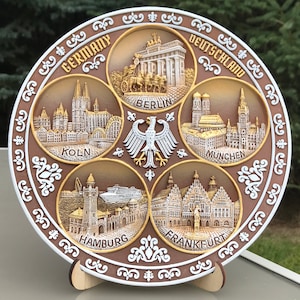 May include: A brown ceramic plate with a white border and gold accents. The plate is divided into six sections, each featuring a different city in Germany: Berlin, Koln, Munchen, Hamburg, Frankfurt, and a section with a white eagle. The plate reads "Germany" and "Deutschland" in gold lettering.