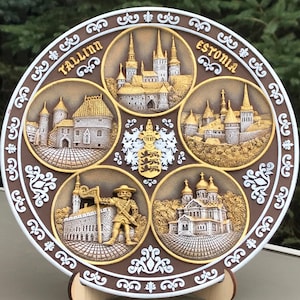 May include: A brown ceramic plate with a gold and white design depicting scenes from Tallinn, Estonia. The plate features the text "Tallinn" and "Estonia" in gold lettering. The plate is on a wooden stand.