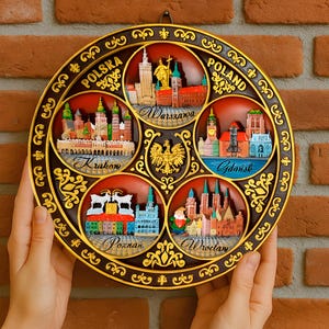 May include: A round decorative plate with a brown background and gold trim. The plate is divided into six sections, each featuring a different city in Poland: Warsaw, Gdansk, Krakow, Poznan, Wroclaw, and a section with the Polish coat of arms. Each section has a 3D image of the city's skyline and the city's name in gold lettering.