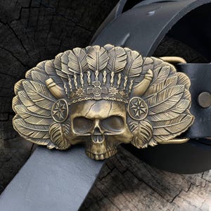 May include: A detailed, antique brass belt buckle featuring a skull wearing a feathered headdress. The buckle is attached to a black leather belt. The skull has intricate details, and the feathers are fanned out on either side.
