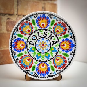 Può includere: Un piatto in ceramica bianca con un design di arte popolare polacca colorato. Il piatto presenta un cerchio centrale con la parola "POLSKA" in lettere nere, circondato da motivi floreali in rosso, blu, giallo e verde.