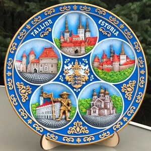 May include: A blue and gold ceramic plate featuring five raised images of buildings in Tallinn, Estonia. The plate has a gold rim with a repeating pattern. The text "Tallinn" and "Estonia" are written in gold on the plate.