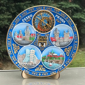 May include: A blue and gold ceramic plate featuring a clock face and images of Prague landmarks. The plate reads "Česko Praha" and depicts the Charles Bridge, the Old Town Square, the Dancing House, and Prague Castle.