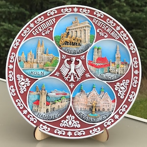 May include: A round, red ceramic plate with a white border and decorative designs. The plate features six raised, circular images of famous German cities: Berlin, Cologne, Munich, Hamburg, Frankfurt, and a German eagle. The plate is titled "Germany" and "Deutschland" in English and German.
