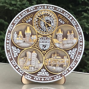May include: A round, decorative plate with a brown background and gold accents. The plate features five images of Prague landmarks, including the Astronomical Clock, Charles Bridge, and the Old Town Square. The plate also includes the words "Česko" and "Praha" in gold lettering.