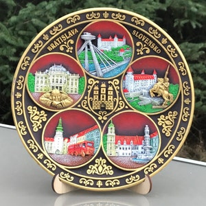 May include: A round, black and gold ceramic plate with a decorative design featuring images of buildings and landmarks in Bratislava, Slovakia. The plate has the words "Bratislava" and "Slovensko" written on it.