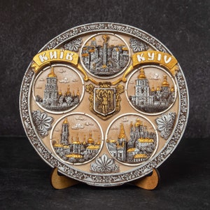 May include: A decorative ceramic plate with a raised design of buildings and the words "КИЇВ" and "KYIV" in gold lettering. The plate is on a wooden stand.