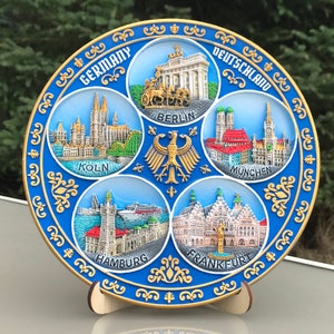 May include: A blue and gold ceramic plate with a decorative border. The plate features images of famous landmarks in Germany, including the Brandenburg Gate, Cologne Cathedral, Munich's Frauenkirche, Hamburg's Elbphilharmonie, and Frankfurt's Römer.