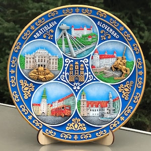 May include: A blue and gold ceramic plate featuring five raised images of landmarks in Bratislava, Slovakia. The plate has a gold rim and a decorative border. The text "BRATISLAVA" and "SLOVENSKO" are written on the plate.