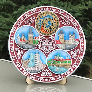 May include: A round ceramic plate with a decorative design featuring images of Prague, Czech Republic. The plate is red, white and gold and has the words "Česko" and "Praha" written on it.