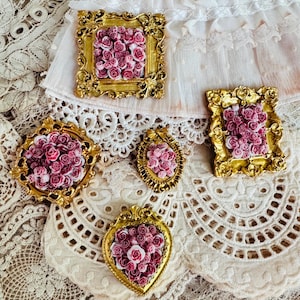 May include: Four decorative wall ornaments featuring ornate gold-colored frames. Each frame contains a cluster of small, three-dimensional pink and white rose-shaped embellishments. The frames vary in shape, including rectangular, oval, and heart-shaped designs.