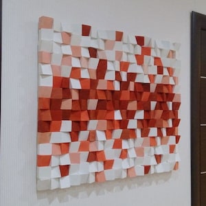 May include: A square, textured wall art piece composed of small, uneven wooden blocks. The blocks are painted in shades of white, peach, and red, creating an abstract, geometric design. The artwork is mounted on a white wall, with dark brown doors on either side.