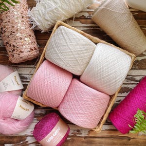 May include: A selection of yarn in different colours and textures. Includes pink, white, and beige yarn cones, plus a basket of pink and white yarn balls. Some yarn is decorated with sequins and glitter.
