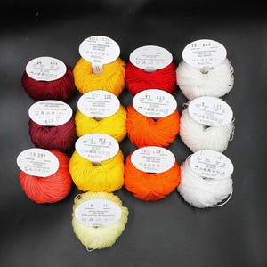 May include: A collection of colorful yarn skeins in various shades of red, yellow, orange, and white. Each skein has a label with text and numbers. The yarn is arranged on a dark surface, creating a visually appealing display for crafting projects.