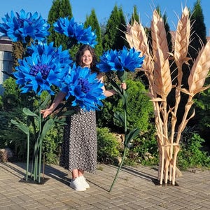 Op de afbeelding: Een persoon poseert met grote, levendige blauwe kunstbloemen en gouden tarwearen. De oversized bloemen hebben gedetailleerde bloemblaadjes en donkerblauwe centra. De tarwearen zijn aan elkaar gebonden. De scène speelt zich buiten af met groen gebladerte op de achtergrond.