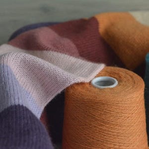 May include: A close-up shot of yarn spools and fabric in various colours. The yarn is in shades of orange, pink, and purple. The fabric is a soft, knitted material with a colour block design. The image highlights the texture of the yarn and fabric.