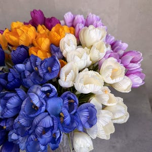 May include: A vibrant bouquet of colorful tulips in shades of blue, yellow, purple, and white. The flowers are tightly clustered, creating a full and textured display. The arrangement is set against a neutral gray background.