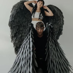 May include: Large, dark grey angel wings with a layered feather design. A person is wearing the wings, with another person standing behind them. The background is a plain white wall.