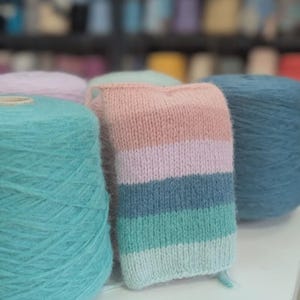 May include: Close-up of a knitted swatch with horizontal stripes in pink, blue, and green. The swatch is next to large spools of turquoise, teal, and lavender yarn. The image suggests a craft or textile theme, with the yarn likely for knitting.
