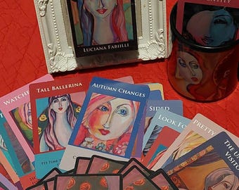 May include: A deck of 44 oracle cards called "Muses of Inspiration" by Luciana Fabilli. The cards feature colorful illustrations of women with unique hairstyles and clothing. The cards are spread out on a red surface with a mug and a framed card.